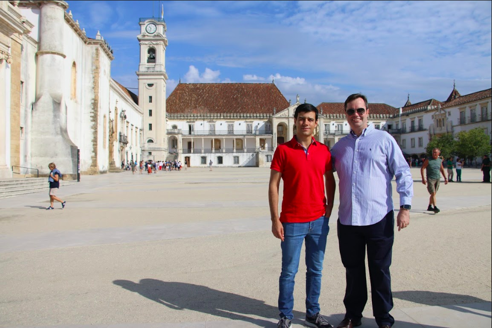 Descrição da imagem: Foto mostra em primeiro plano o professor Joel Arrais ao lado do professor Fabiano Papaiz, os dois estão em pé posando para a foto. Ao fundo vemos um prédio da Universidade de Coimbra, ele é branco e apresenta características antigas. 