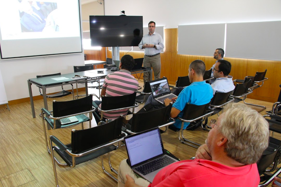 Descrição da imagem: ao fundo da imagem o professor Fabiano Papaiz apresenta slides em uma tela de projeção grande. Cinco homens estão sentados à sua frente assistindo a apresentação, dois deles estão com notebooks no colo. 