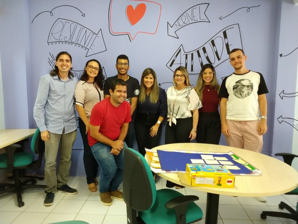 Descrição da imagem: Oito participantes da oficina posam para foto ao final da atividade. Em primeiro plano, uma mesa redonda marrom com materiais em cima. O fundo da foto é uma parede onde estão escritas as palavras Resiliente, Incrível e Atitude. Também existe um coração pintado na mesma parede. 